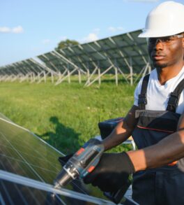 African american man in safety helmet and glasses tighten nuts on solar panels with screwdriver. Competent technician using tools while performing service work on station.